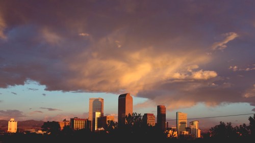 City skyline at sunset with dramatic clouds and a warm glow illuminating the buildings.