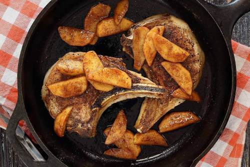 Two pork chops cooked in a skillet, topped with sliced apples, on a checkered tablecloth.
