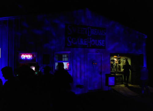A spooky scare house at night, illuminated by blue lights and an "Open" sign, with people gathered outside.