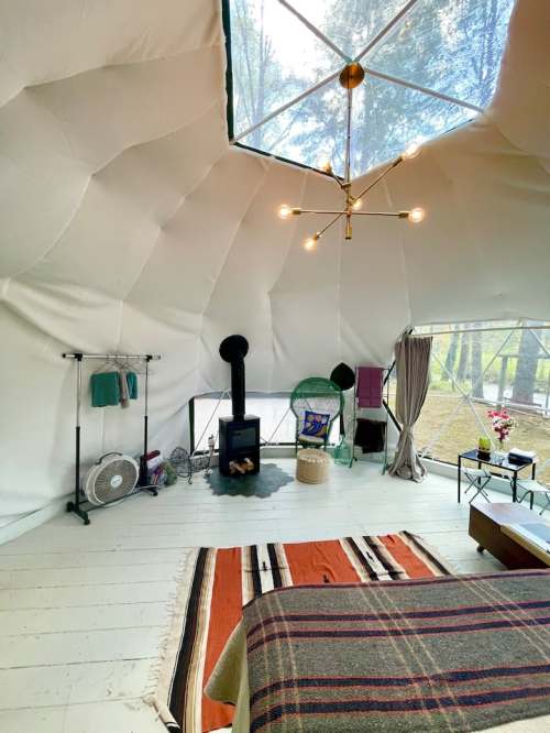Cozy interior of a geodesic dome with a bed, stove, and decorative elements, surrounded by natural light.