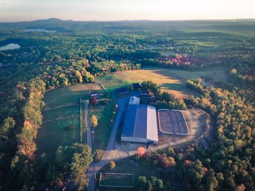 Aerial view of a large green landscape with a barn and riding arena, surrounded by trees and hills in the background.