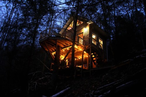 A cozy treehouse illuminated at night, nestled among dark trees with warm lights glowing from its windows and deck.