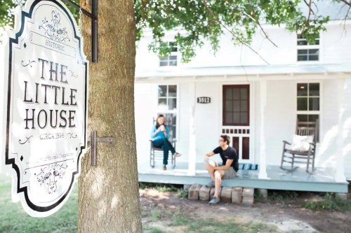 A historic sign for "The Little House" with two people relaxing on the porch of a white house.
