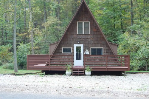 A cozy A-frame cabin surrounded by trees, featuring a front deck and gravel pathway.