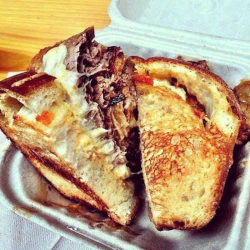 A close-up of a sandwich with layers of beef and melted cheese, served in a takeout container.