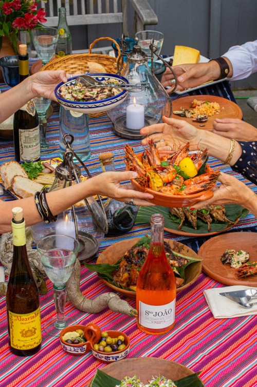 A vibrant table setting with various dishes, including shrimp, wine, and colorful textiles, as people share food.
