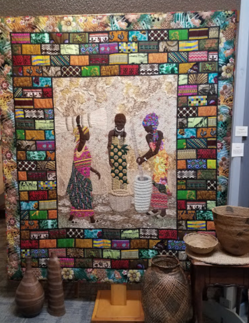 A vibrant quilt featuring three women in traditional attire, surrounded by a colorful patterned border and woven baskets.