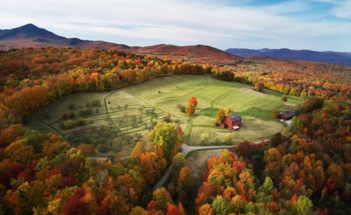 Aerial view of a vibrant autumn landscape with colorful trees, a green field, and a red barn surrounded by mountains.