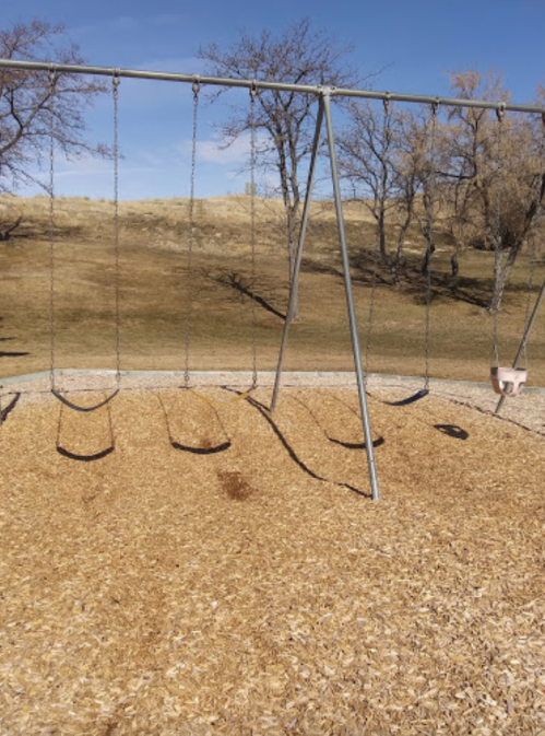 Empty swings on a playground, with a grassy hill and trees in the background under a clear blue sky.