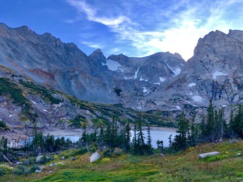 A serene mountain landscape with rugged peaks, a calm lake, and lush greenery under a blue sky.