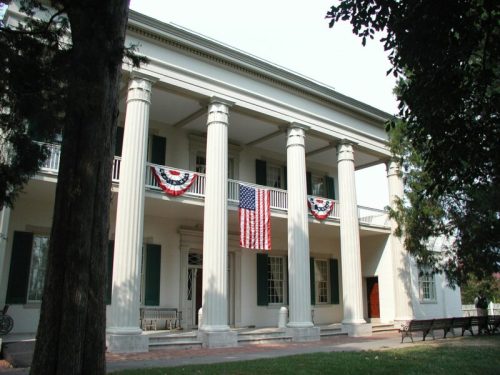 Historic building with large columns, American flag, and patriotic bunting, surrounded by trees.
