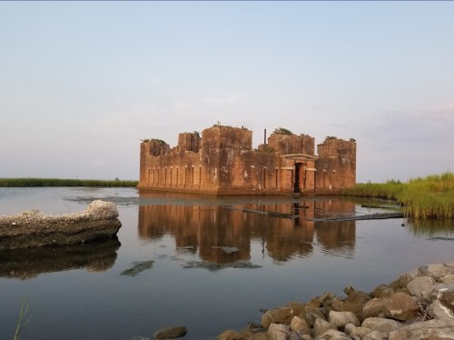 A weathered brick structure stands in calm water, surrounded by greenery and a rocky shoreline under a clear sky.