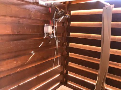 A rustic wooden shower with a handheld showerhead and natural light streaming through slatted walls.