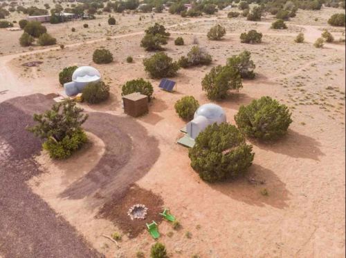 Aerial view of a desert campsite with two domes, a solar panel, a fire pit, and green chairs among sparse vegetation.