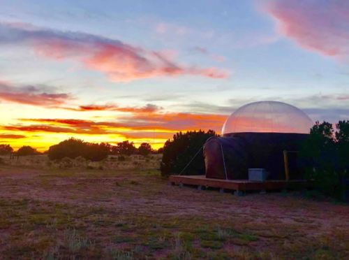 A dome-shaped structure against a vibrant sunset, surrounded by sparse vegetation in a desert landscape.