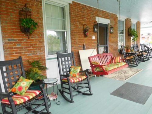 A cozy porch with rocking chairs, colorful cushions, and potted plants against a brick wall.