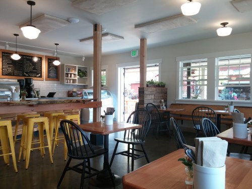 Bright café interior with wooden tables, black chairs, and a counter displaying a menu. Large windows let in natural light.