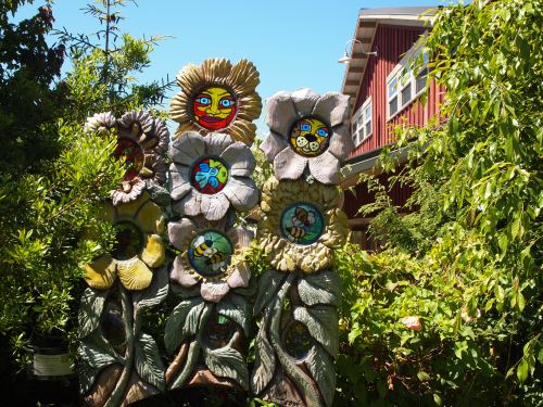 Colorful, whimsical flower sculptures with painted faces, surrounded by lush greenery and a red building in the background.