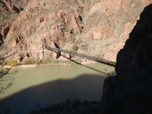 A wooden bridge spans a river, surrounded by rocky cliffs and rugged terrain under bright sunlight.