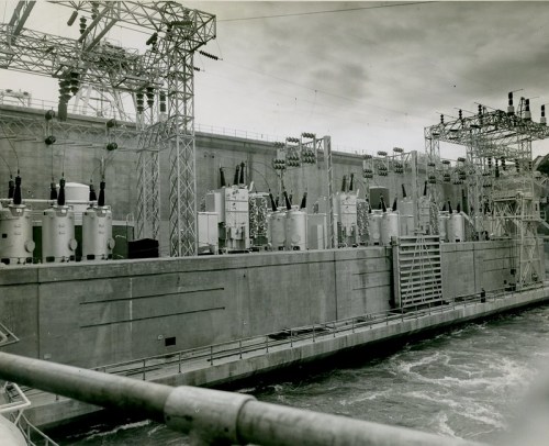 Black and white image of a large hydroelectric power station with transformers and electrical equipment along a dam.