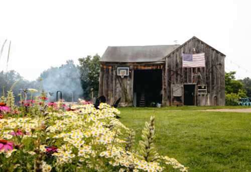 A rustic barn with an American flag, surrounded by colorful flowers and a hint of smoke in the background.