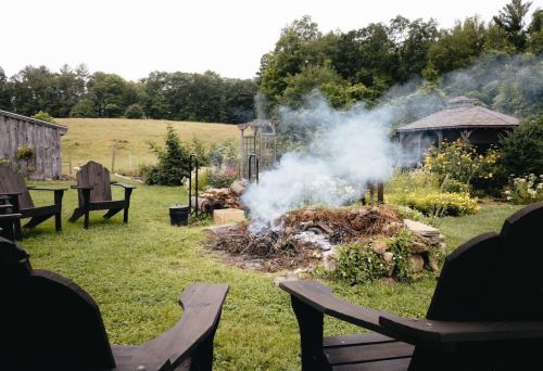 A serene outdoor scene with smoke rising from a fire pit, surrounded by chairs and lush greenery in the background.