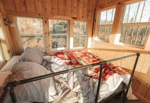 Cozy bedroom with a bed covered in blankets, surrounded by large windows and wooden walls, letting in warm sunlight.