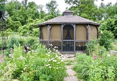 A round, wooden garden shed surrounded by lush greenery and colorful flowers, with a windmill in the background.