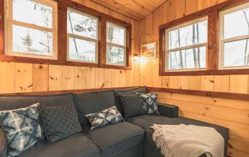 Cozy cabin interior with a sectional sofa, decorative pillows, and large windows overlooking a wooded area.