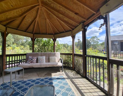 A cozy gazebo with a cushioned bench, table, and a view of a garden under a blue sky with scattered clouds.