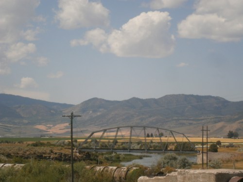A metal bridge spans a river, surrounded by rolling hills and fields under a partly cloudy sky.