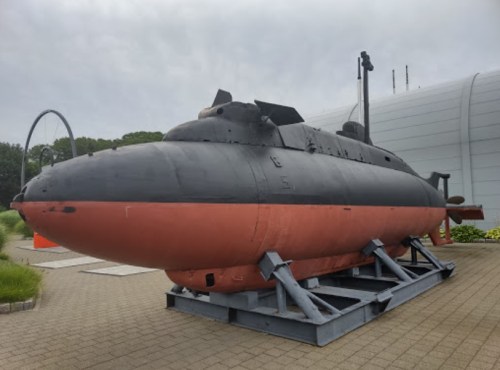 A black and red submarine displayed on a stand, set against a cloudy sky and surrounded by greenery.