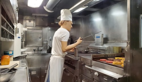 A chef in a white hat cooks colorful burgers on a grill in a stainless steel kitchen.
