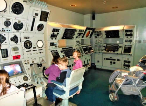 Children interact with vintage control panels in a retro-themed room, with a stroller in the background.