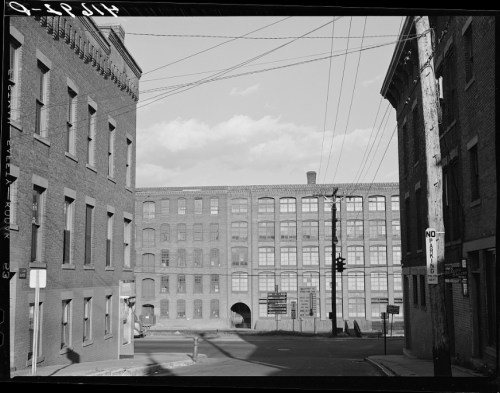 A view of a street corner with brick buildings and a large factory in the background, under a clear sky.