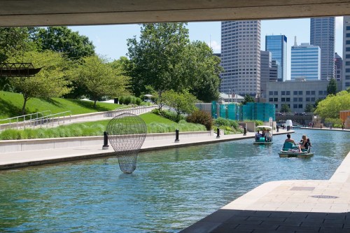 A serene urban canal scene with boats, greenery, and city skyscrapers in the background under a sunny sky.