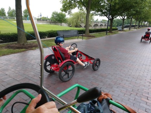 A child in a red go-kart rides along a paved path in a park, wearing a helmet and enjoying the outdoors.