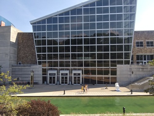 A modern glass building with a large entrance, surrounded by greenery and a walkway along a body of water.