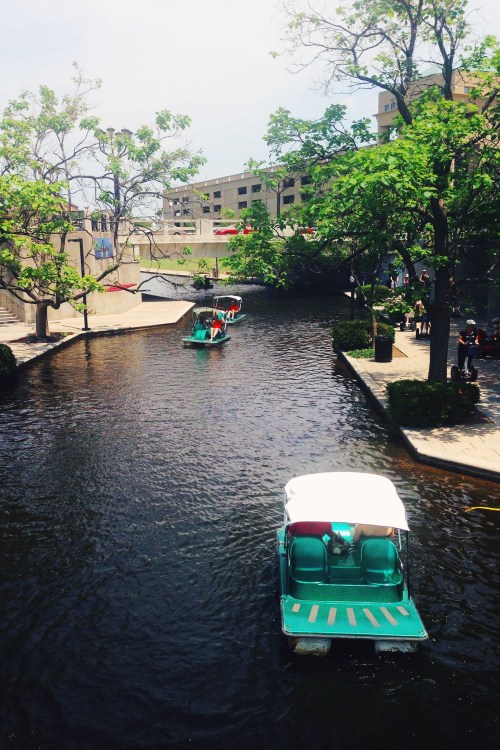 A serene canal scene with small boats gliding through the water, surrounded by trees and urban buildings.