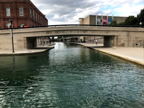 A view of a canal with a stone bridge, surrounded by buildings and people walking along the pathways.