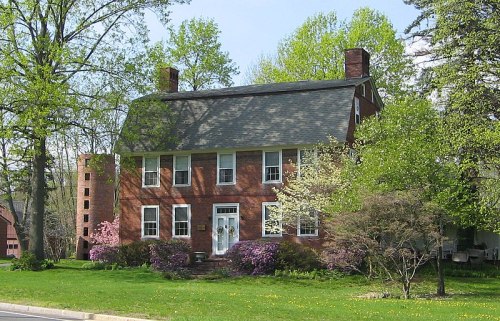 A two-story brick house surrounded by green trees and blooming flowers in a sunny setting.