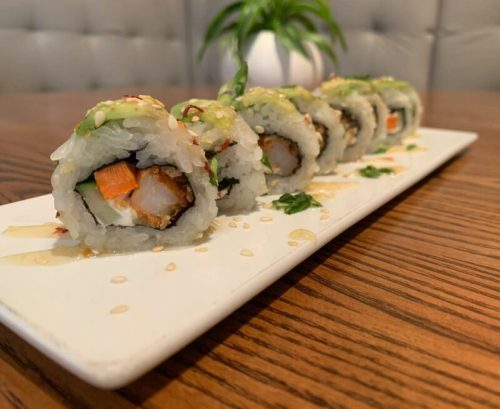 A plate of sushi rolls with shrimp, avocado, and vegetables, drizzled with sauce, on a wooden table.