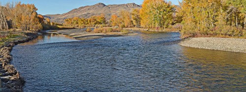 A serene river winds through a landscape of autumn trees and mountains under a clear blue sky.