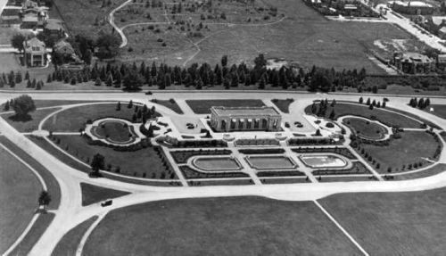 Aerial view of a grand building surrounded by landscaped gardens and pathways, with nearby residential areas.