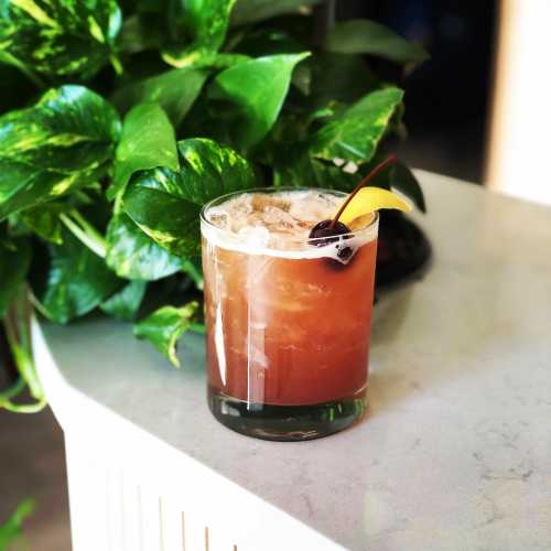 A refreshing cocktail with ice, garnished with a cherry, sits on a marble surface next to lush green foliage.