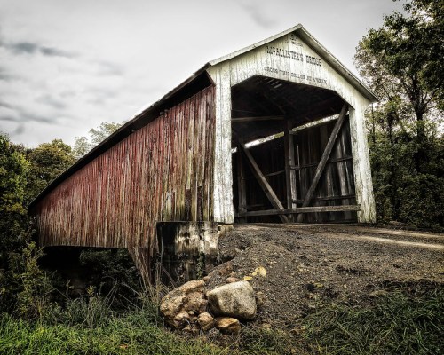 A weathered covered bridge with a wooden structure, surrounded by greenery and a gravel path.
