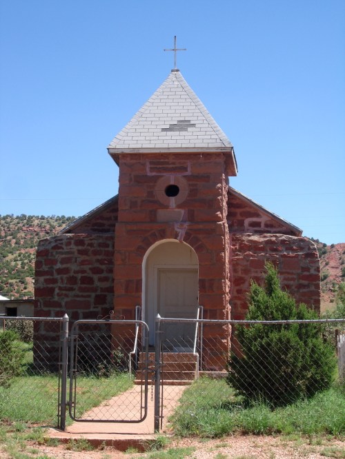 A small stone church with a cross on top, surrounded by a fence and greenery, set against a clear blue sky.