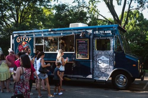 A blue food truck named "Gypsy" serves crepes, with a line of customers waiting outside in a park setting.
