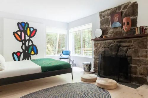 A modern bedroom with a stone fireplace, colorful wall art, and large windows overlooking a forest.