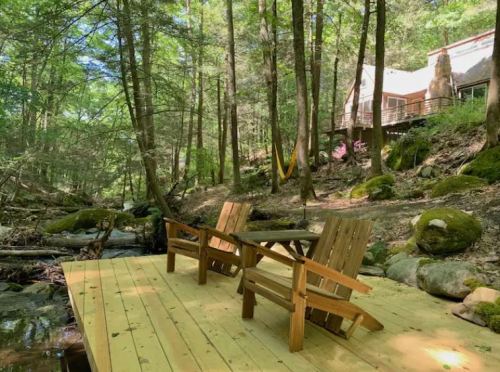 A wooden deck by a stream in a forest, featuring two chairs and a cozy cabin in the background.